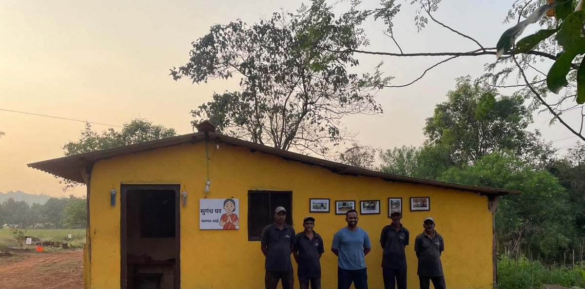 People standing outside the yellow Sugandha Ghar building beneath trees in Tithavli, Konkan.