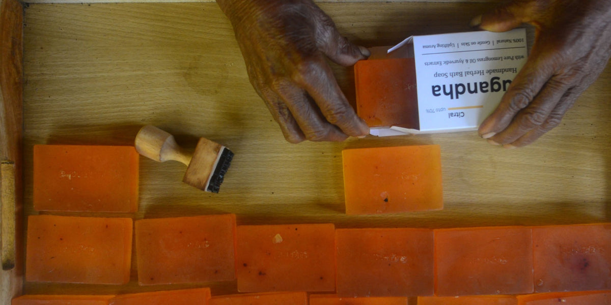 Hands packing a Sugandha soap at a wooden worktable beside unpacked soap bars.